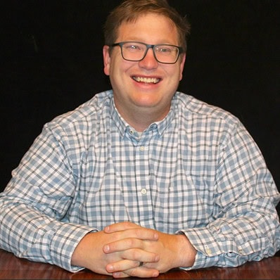 Christopher Kolks smiling at a desk, wearing glasses and a checkered shirt.