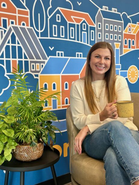 Kaili McCool smiling with a mug in front of colorful cityscape mural.