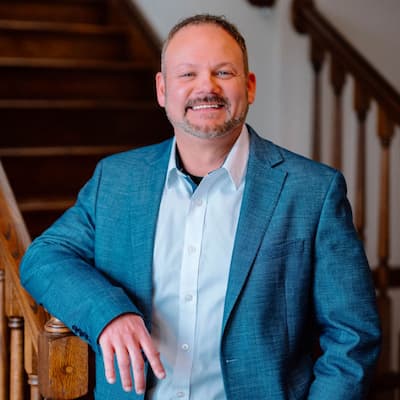 Dan Bihn smiling in a blue suit, standing by a wooden staircase.