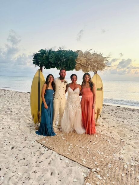 Beach wedding celebration with four people standing in front of surfboards on the sand.
